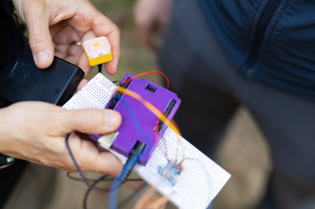 A person offscreen holds sensor components into the frame: a small bright purple box, a black box, a white breadboard, a small yellow box with an LED turned on in the middle, while the person on the right holds a few of the wires against a reusable water bottle.