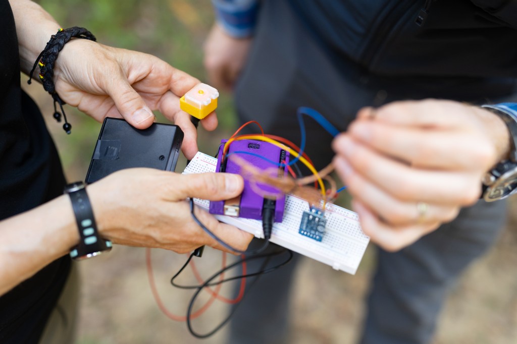 Two people's hands hold sensor components into the frame: the person on the left holds a small bright purple box, a black box, a white breadboard, a small yellow box with an LED turned on in the middle, while the person on the right holds a few of the wires coming off of the other components.