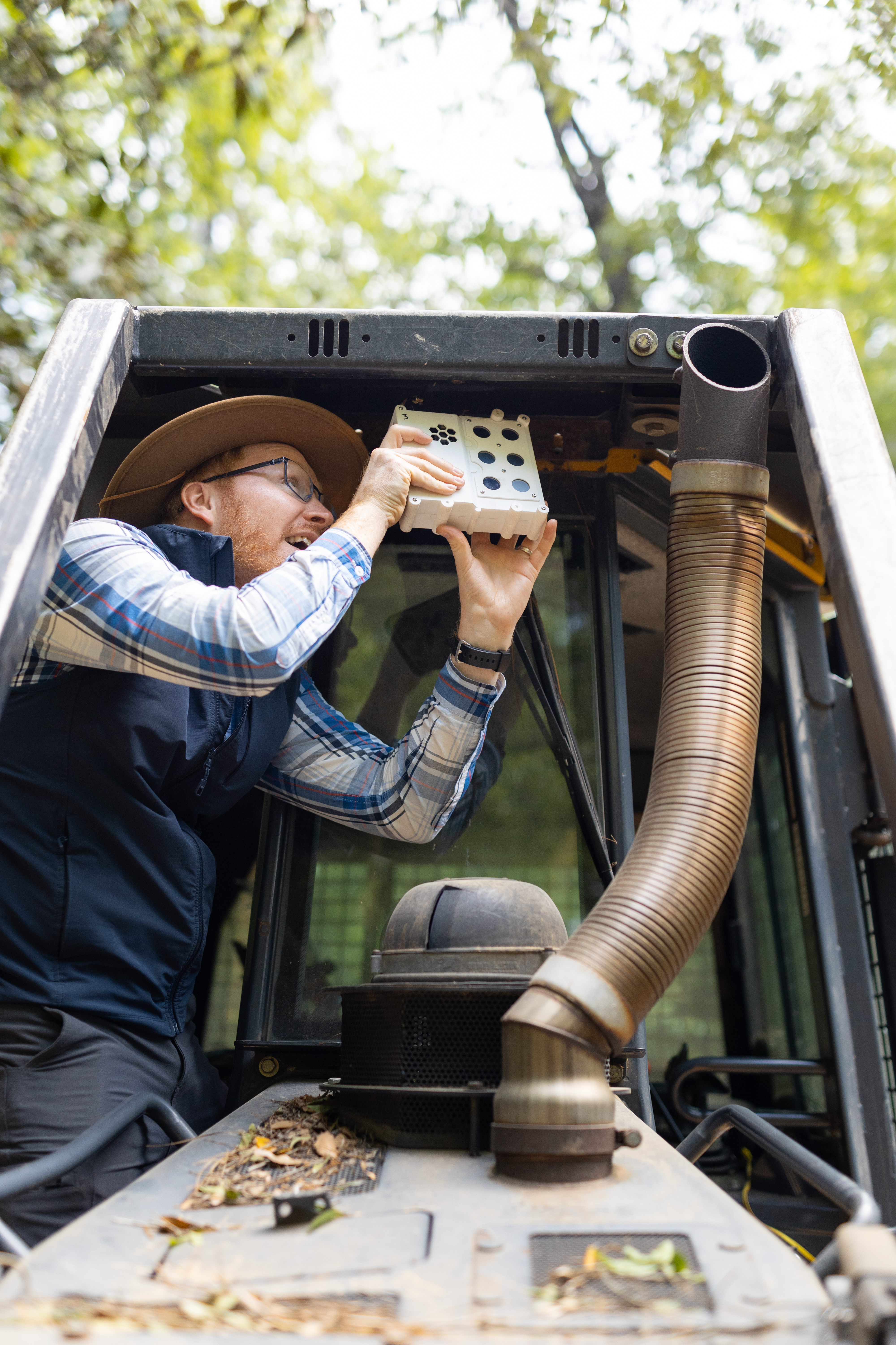 A man in dark gray jeans, a blue checkered long-sleeve, black vest, and straw hat stands on the tread of a black and yellow bulldozer, holding a white sensor box the size of a lunchbox up to the roof in front of the cab. In the background is a green forest.