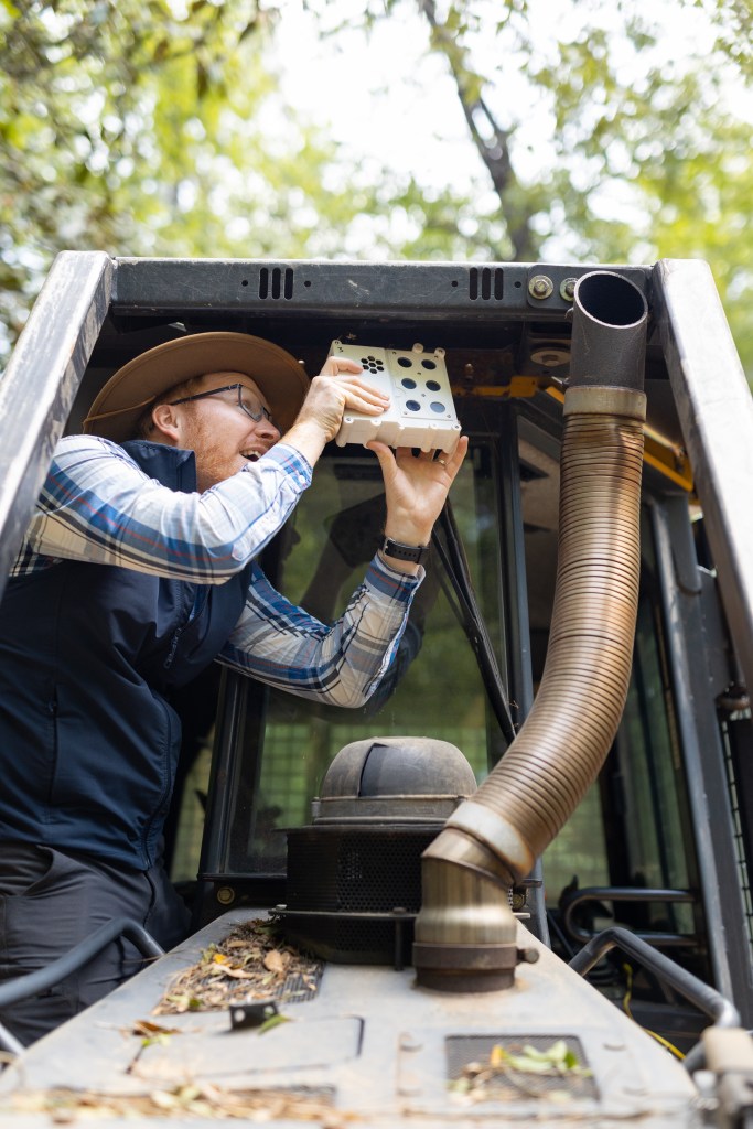 A man in dark gray jeans, a blue checkered long-sleeve, black vest, and straw hat stands on the tread of a black and yellow bulldozer, holding a white sensor box the size of a lunchbox up to the roof in front of the cab. In the background is a green forest.