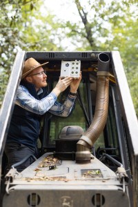 A man in dark gray jeans, a blue checkered long-sleeve, black vest, and straw hat stands on the tread of a black and yellow bulldozer, holding a white sensor box the size of a lunchbox up to the roof in front of the cab. In the background is a green forest.