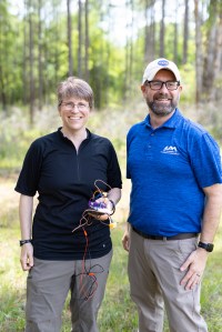Two people stand in a green forest smiling at the camera, with the frame cut off at their knees. On the left is a woman in a black t-shirt, olive green pants, and wire-rimmed glasses, holding a jumble of wires. On the right is a man in a bright blue t shirt, tan pants, a white baseball cap, black glasses and a short beard.