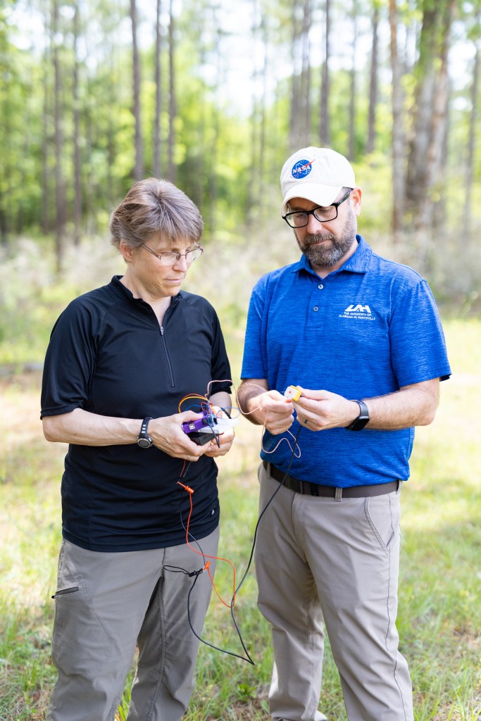 Two people stand in a green forest looking down at the jumble of wires in their hands, with the frame cut off at their knees. On the left is a woman in a black t-shirt, olive green pants, and wire-rimmed glasses. On the right is a man in a bright blue t shirt, tan pants, a white baseball cap, black glasses and a short beard.