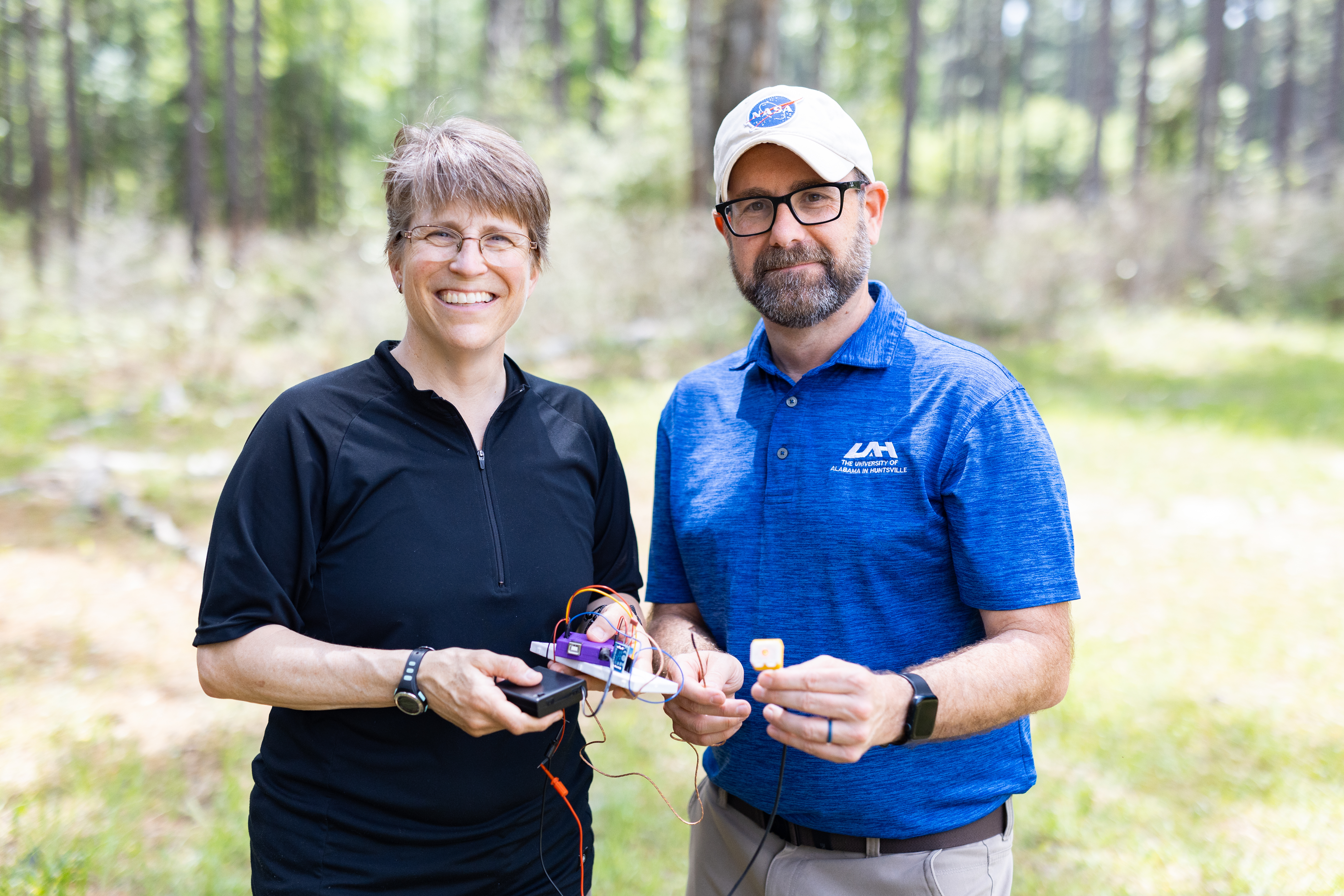Two people stand in a green forest smiling at the camera and holding a jumble of wires and plastic boxes, with the frame cut off at their waist. On the left is a woman in a black t-shirt, olive green pants, and wire-rimmed glasses. On the right is a man in a bright blue t shirt, tan pants, a white baseball cap, black glasses and a short beard.