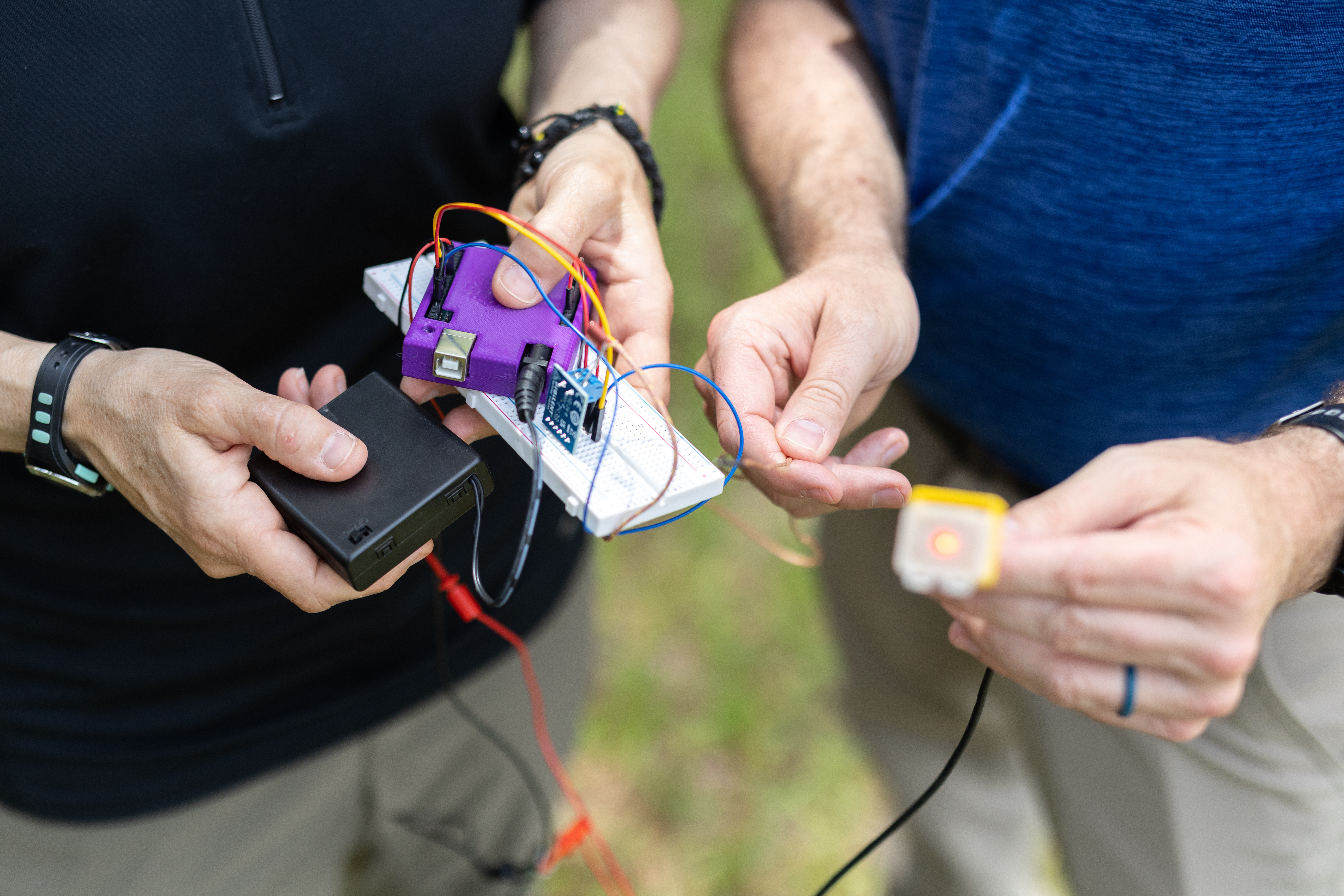Two people's hands hold sensor components into the frame: the person on the left holds a small black box, bright purple box, and white breadboard, while the person on the right holds a small yellow box with an LED turned on in the middle. All of the boxes are connected by a jumble of wires.