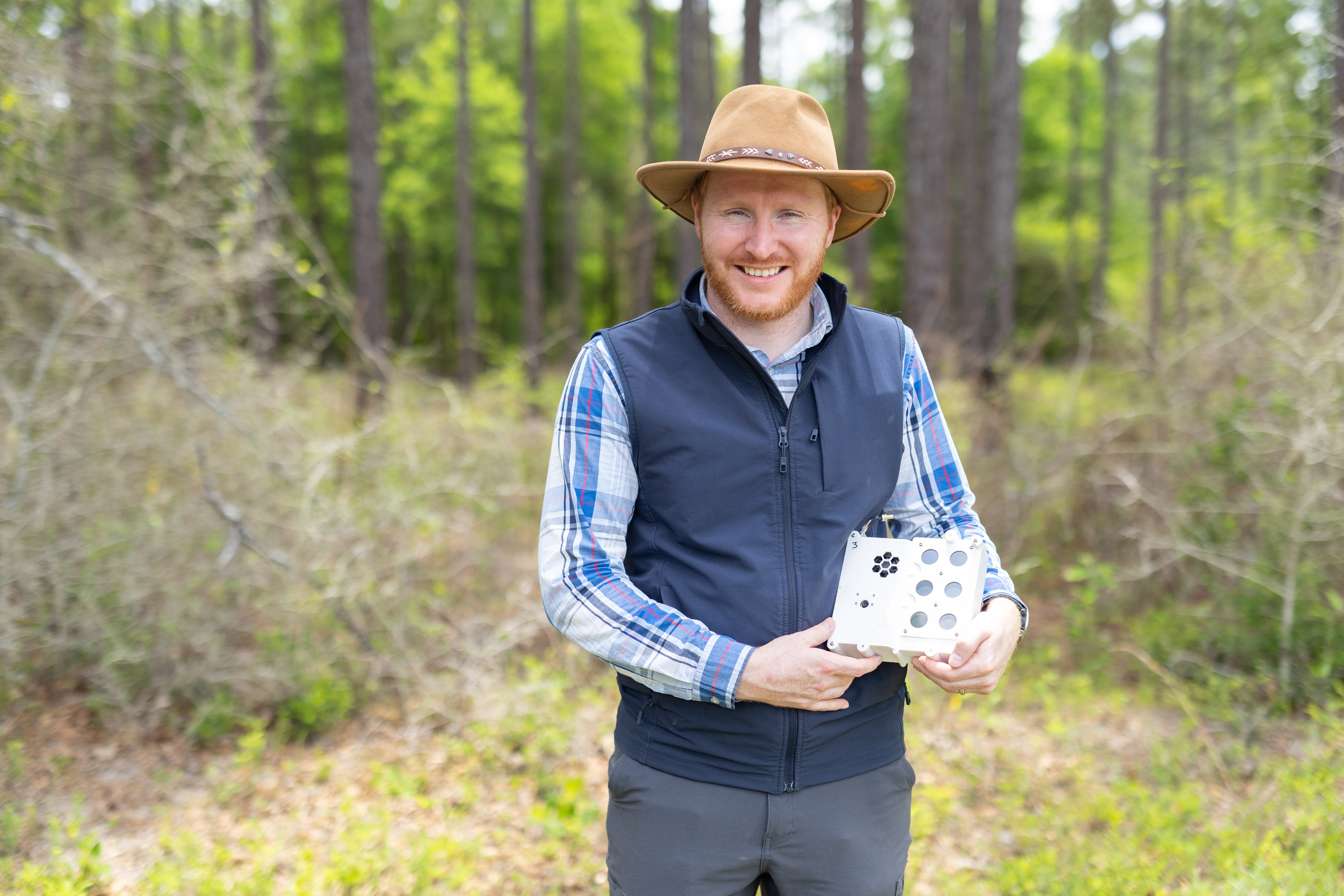 A man in gray pants, a checkered blue long sleeve, black vest, tan hat, and red beard stands in a forest smiling at the camera. He holds a white box the size of a lunchbox on his left hip.