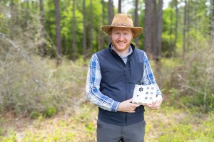 A man in gray pants, a checkered blue long sleeve, black vest, tan hat, and red beard stands in a forest smiling at the camera. He holds a white box the size of a lunchbox on his left hip.