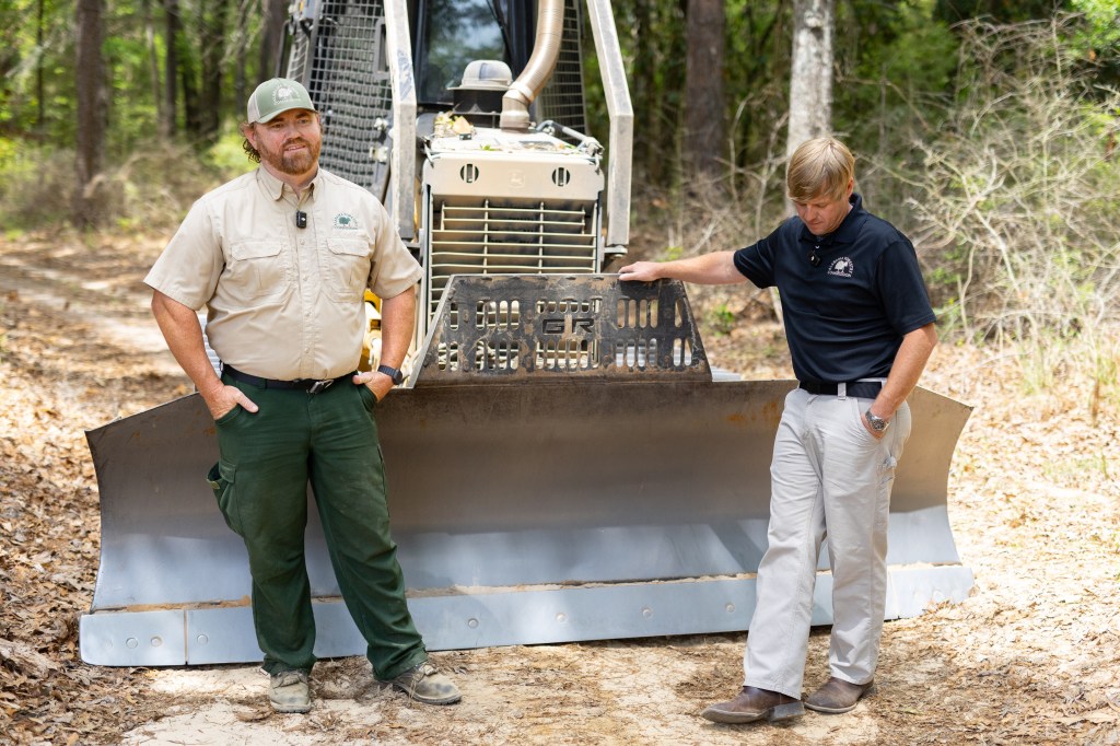 Two men stand in front of a bulldozer blade, on a dirt road in a forest. The man on the left is wearing green pants, a tan t-shirt, and a baseball cap, and standing with his hands in his pockets. The man on the right is wearing a black t-shirt and tan pants, and stands with one hand on the dozer and looking down at the ground.