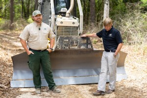 Two men stand in front of a bulldozer blade, on a dirt road in a forest. The man on the left is wearing green pants, a tan t-shirt, and a baseball cap, and standing with his hands in his pockets. The man on the right is wearing a black t-shirt and tan pants, and stands with one hand on the dozer and looking down at the ground.