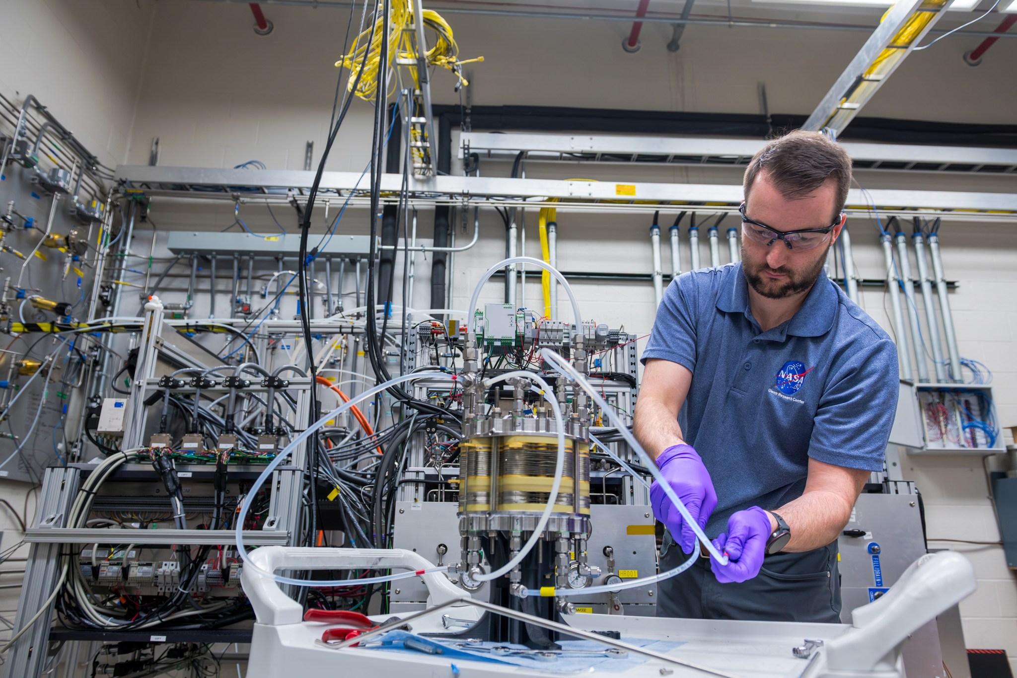 A man wearing a blue NASA polo, purple gloves, and safety glasses adjusts a small tube connected to a silver and yellow cylinder inside the Fuel Cell Testing Laboratory. Behind him are numerous other tubes, wires, and sensors connected to a large system and the lab’s wall.
