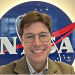 A white man with short brown hair is wearing wire rim glasses, a plaid collared shirt, and tan blazer. He is standing in front of the NASA logo, and the shot cuts off just below the shoulders.