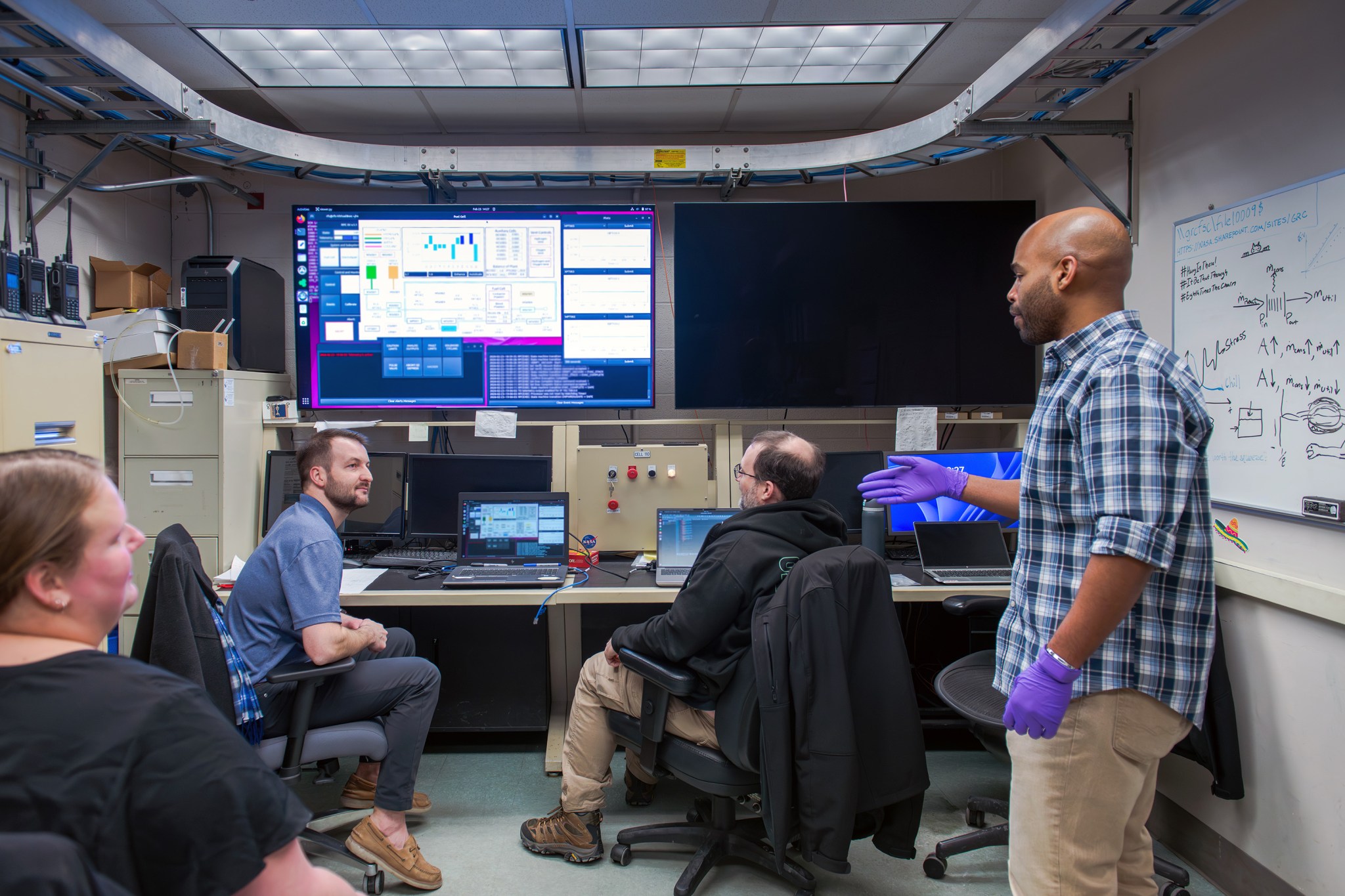 Two researchers stand in a control room and converse with two other researchers sitting in chairs behind colorful monitors and laptops showing testing data. A whiteboard with equations is shown to their right.