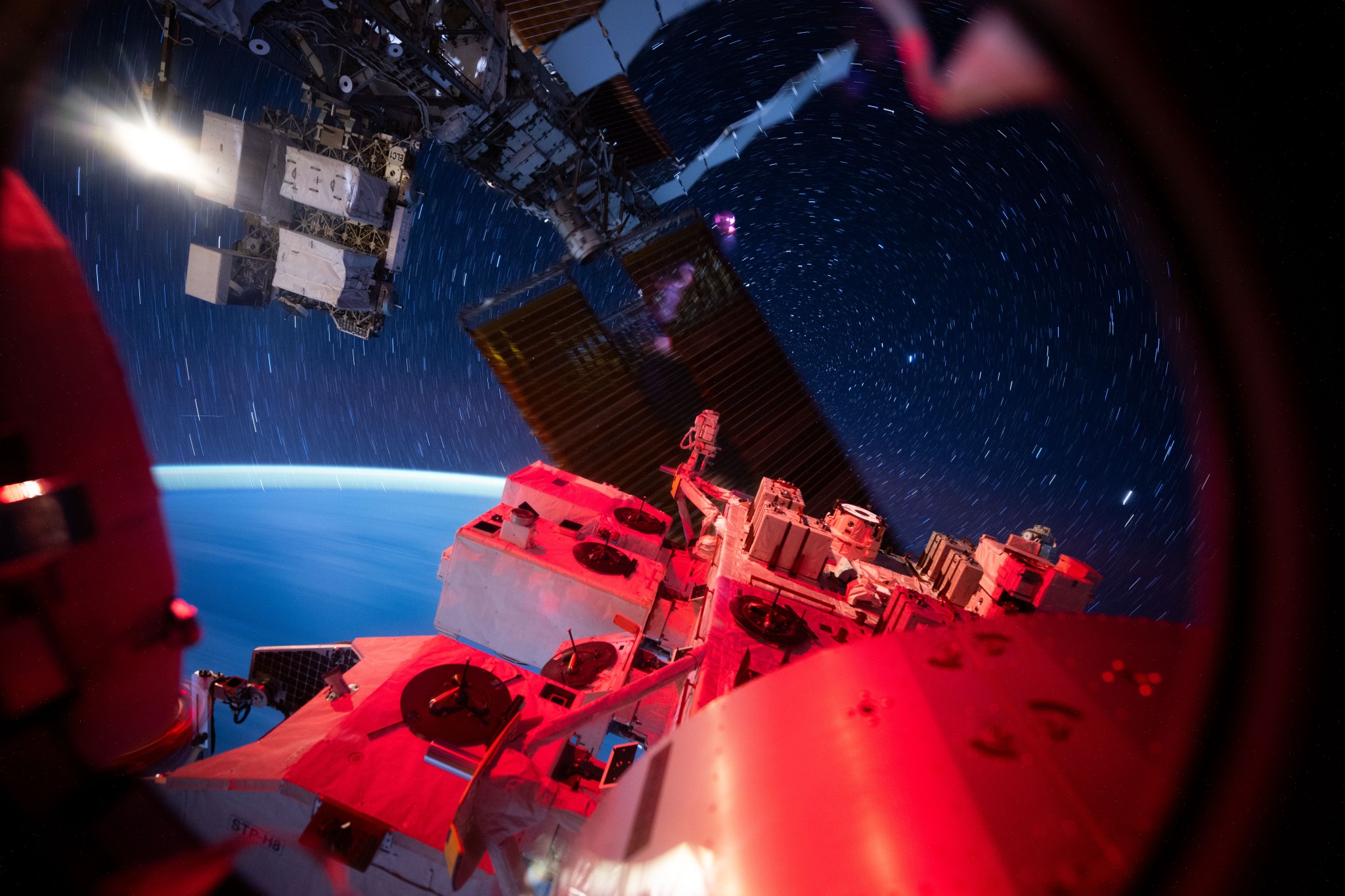 View from the International Space Station showing its illuminated exterior hardware in red light against a backdrop of deep space. Star trails form circular streaks across the sky due to the station’s motion, while Earth’s blue curvature glows softly at the horizon.