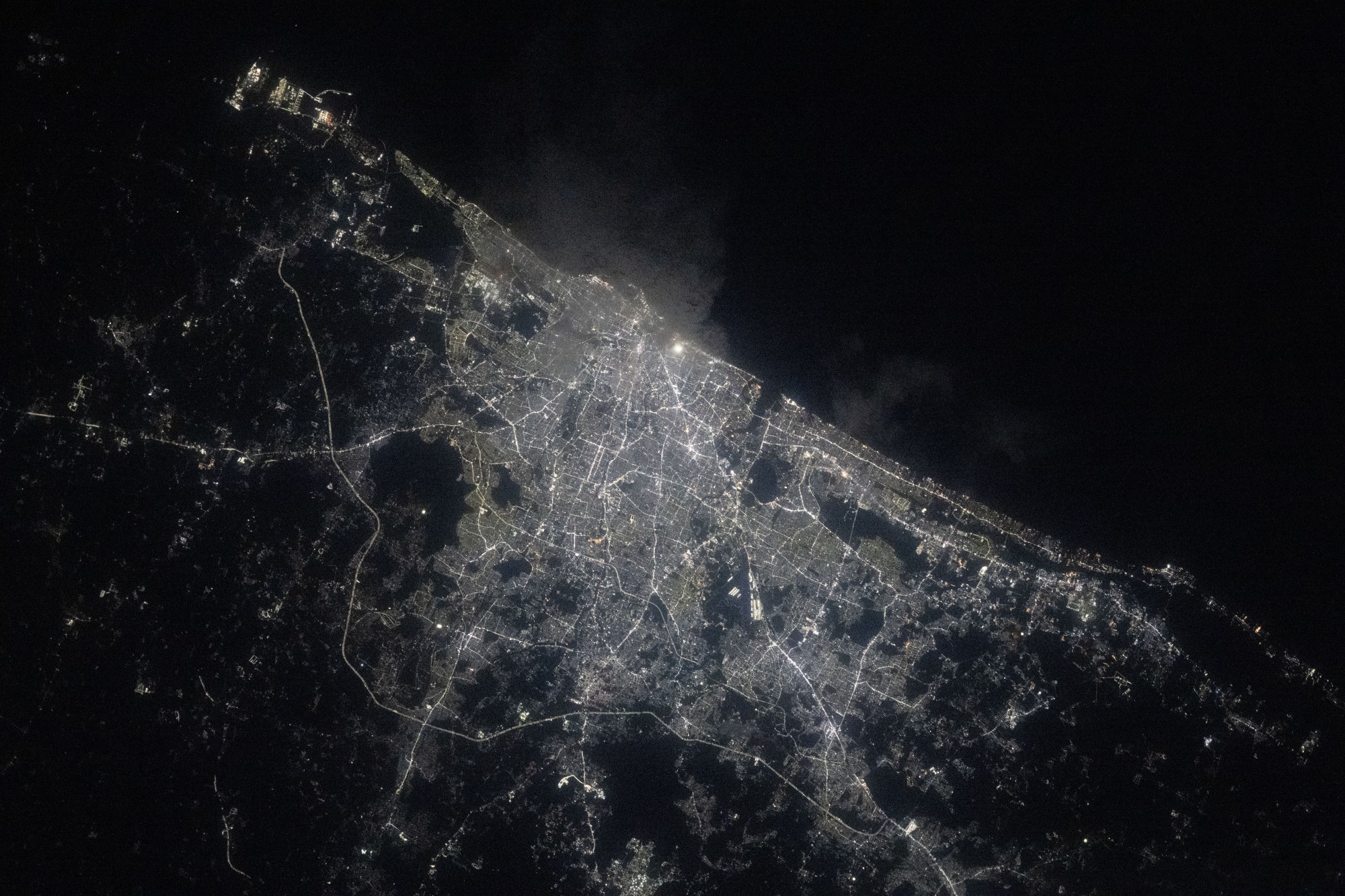 A view of Chennai at night from the International Space Station shows a sprawling city along the water (upper right). City lights make the city shine while also highlighting roadways. The surrounding area is mostly dark.
