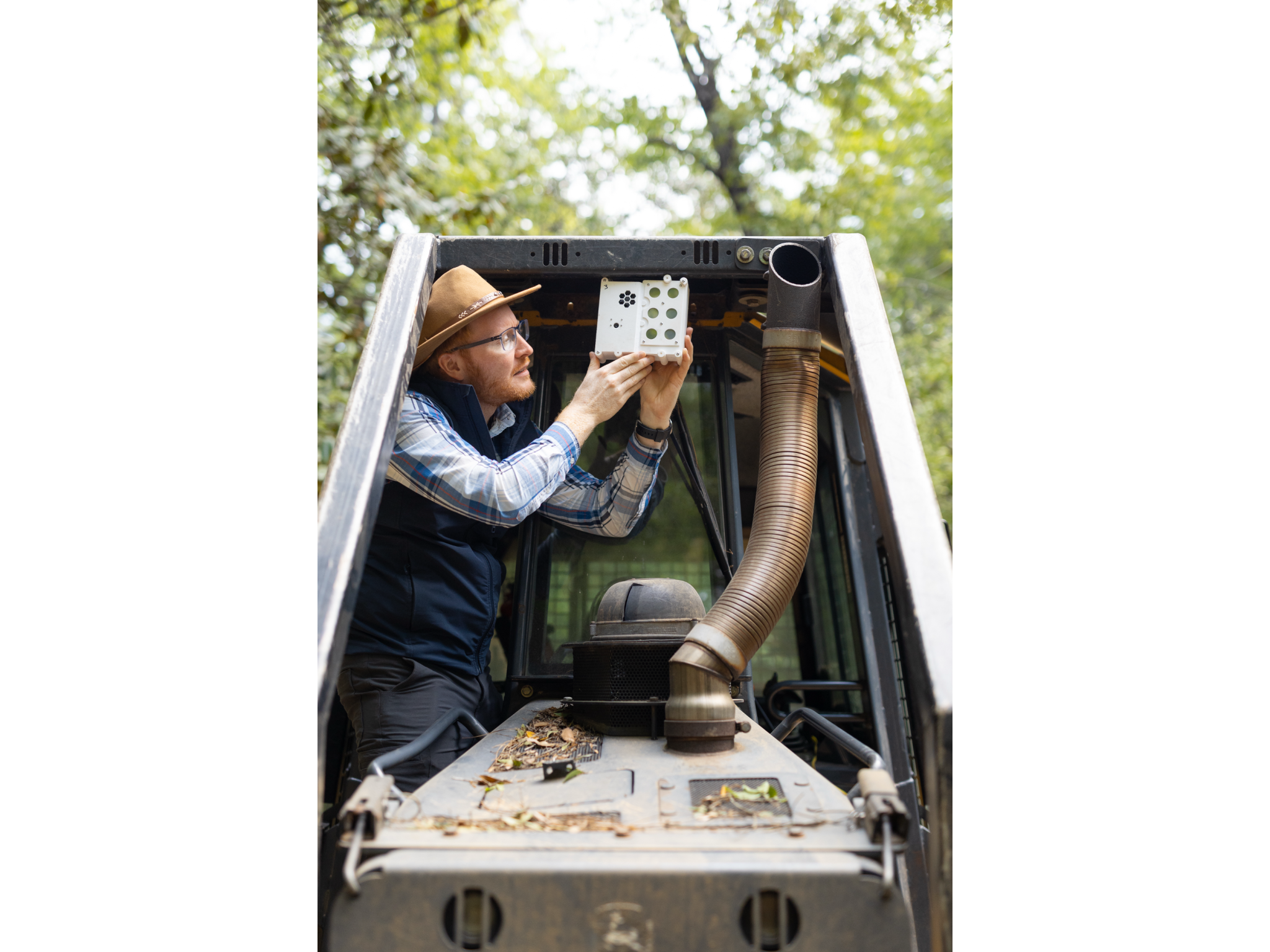 A man in dark gray jeans, a blue checkered long-sleeve, black vest, and straw hat stands on the tread of a black and yellow bulldozer, holding a white sensor box the size of a lunchbox up to the roof in front of the cab. In the background is a green forest.
