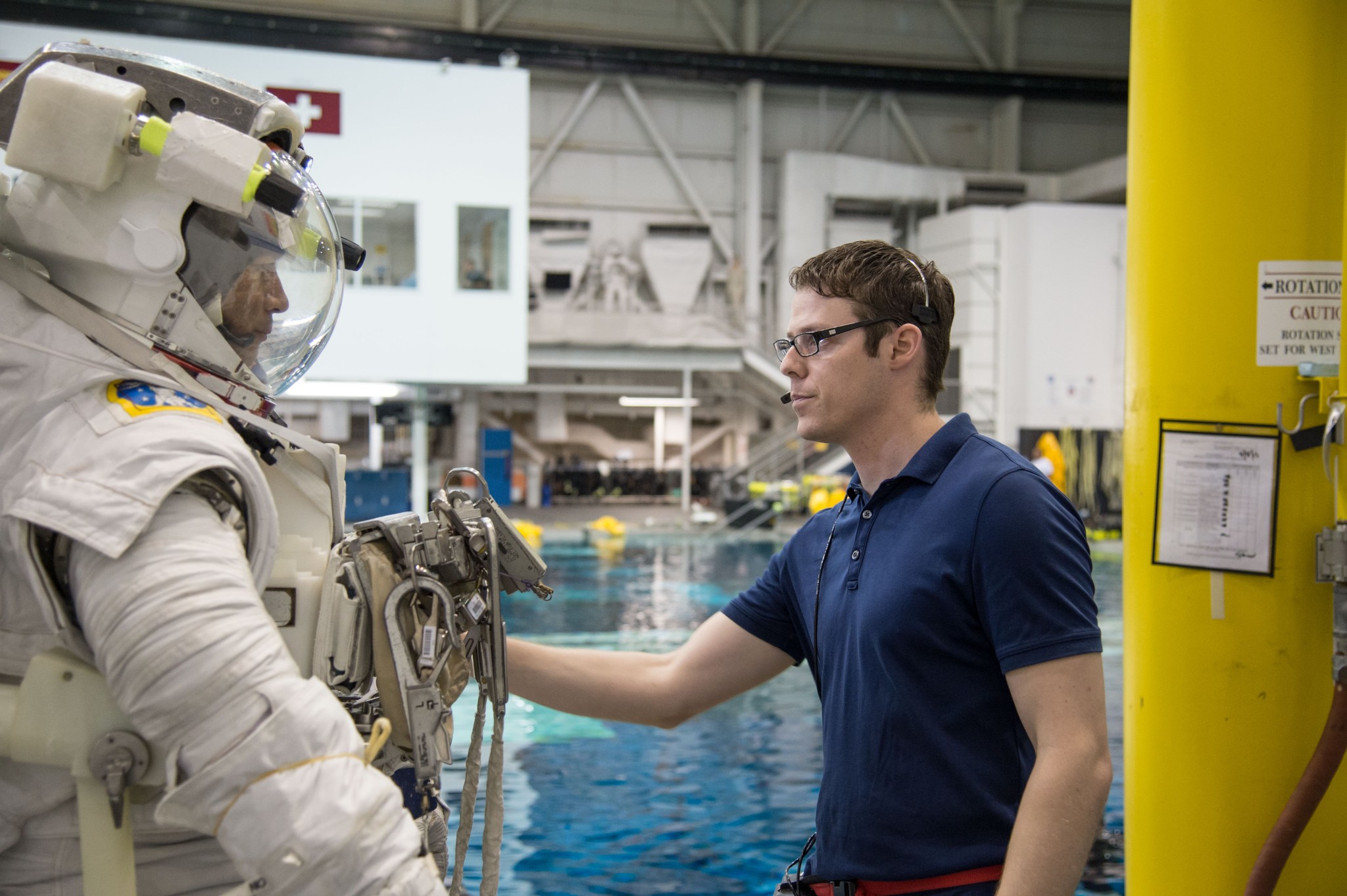A man wearing a blue polo shirt, glasses, and a headset speaks to a suited astronaut on the side of a large pool.