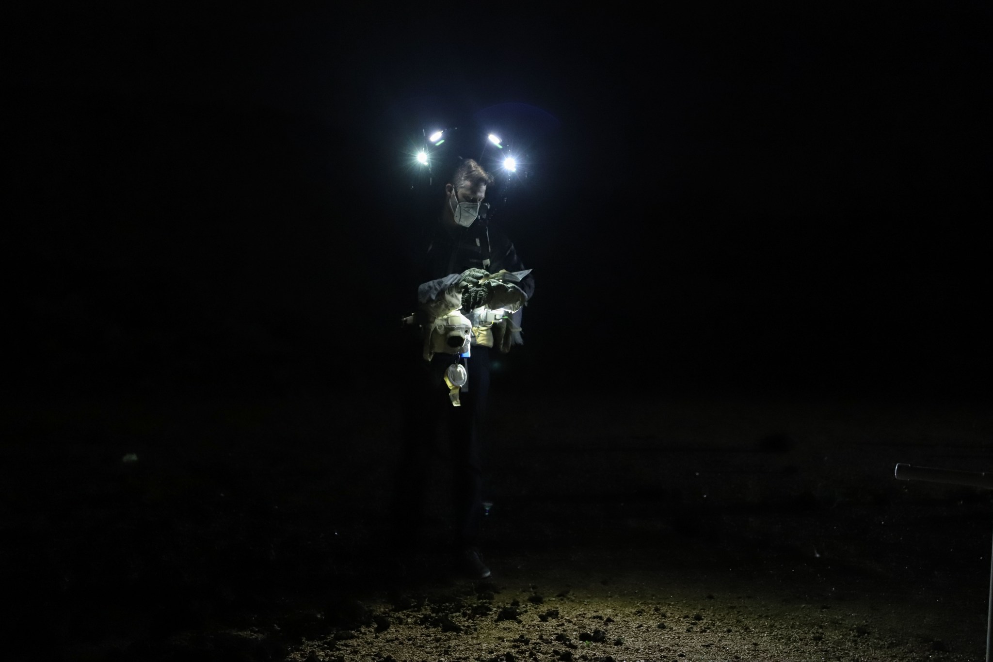 A man wearing an N95 mask, spacesuit gloves, and headlamps studies a booklet of rock samples in the dark.