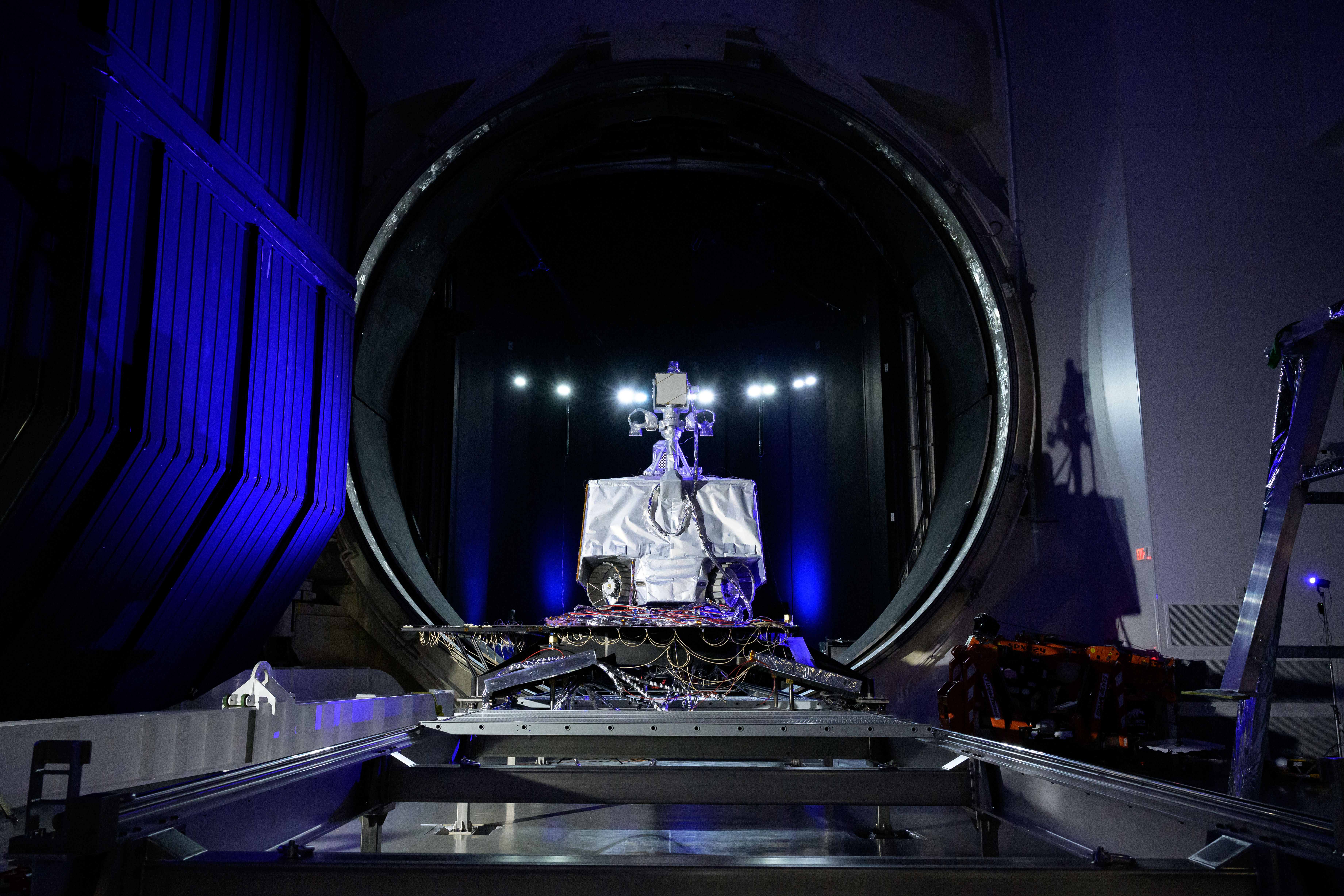NASA’s VIPER rover sits on a testing platform in front of the large circular entrance to Chamber A, a thermal-vacuum test facility at Johnson Space Center. Bright lights illuminate the rover and surrounding equipment inside the dark testing chamber environment.
