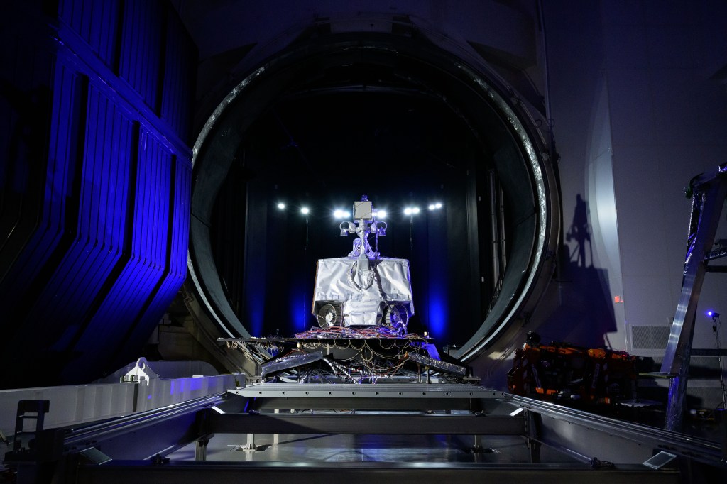 NASA’s VIPER rover sits on a testing platform in front of the large circular entrance to Chamber A, a thermal-vacuum test facility at Johnson Space Center. Bright lights illuminate the rover and surrounding equipment inside the dark testing chamber environment.