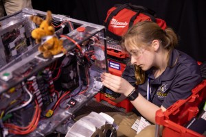 A student holds onto the edge of the rectangular body of a prototype lunar robot as she works on it. The rover has a rectangular body, which has multicolored attached to it. There is a small stuffed moose sitting on top of the rover.