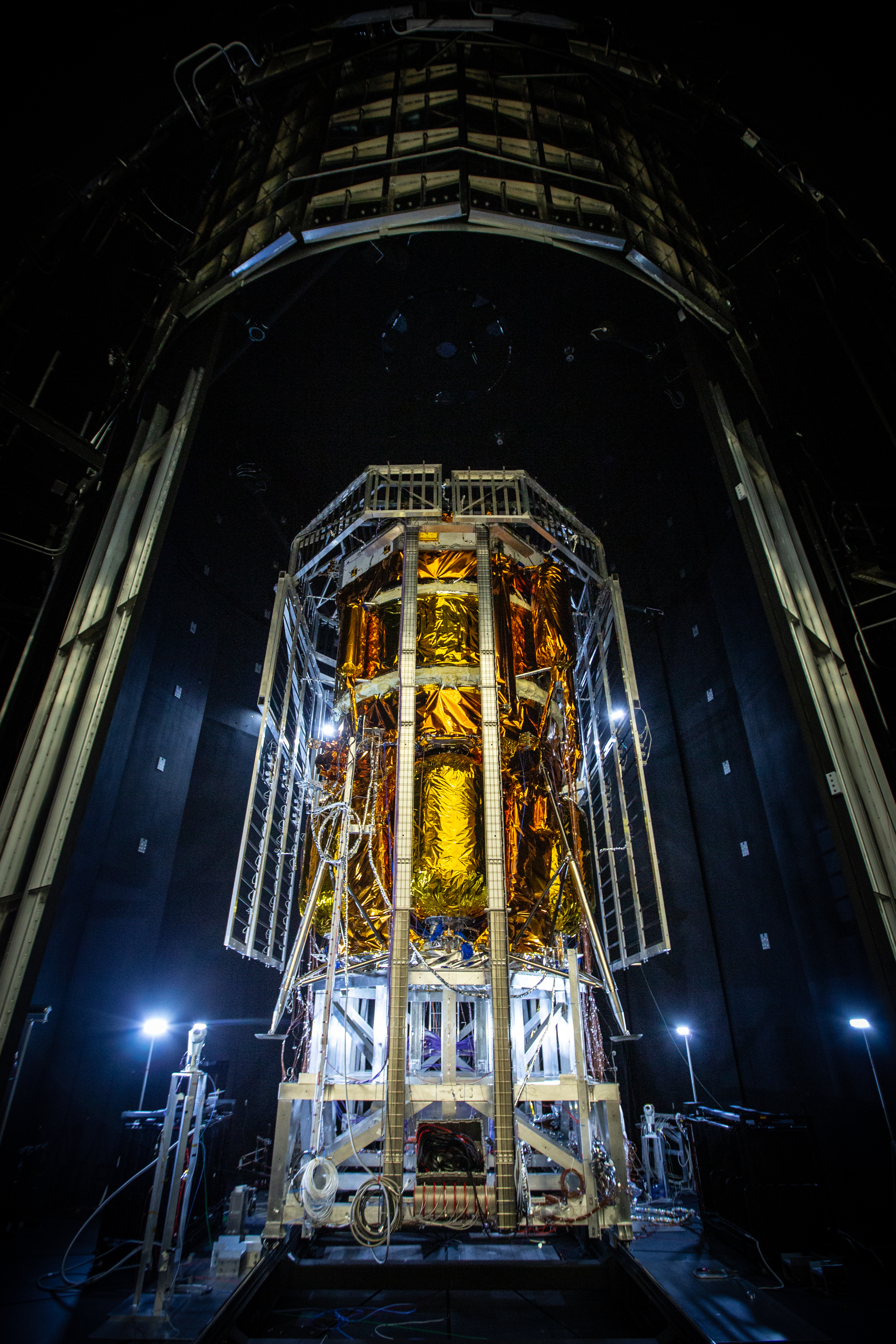 Blue Origin’s Blue Moon Mark 1 lunar lander, covered in gold thermal insulation, stands inside a dark testing chamber surrounded by support structures, cables, and lighting equipment.