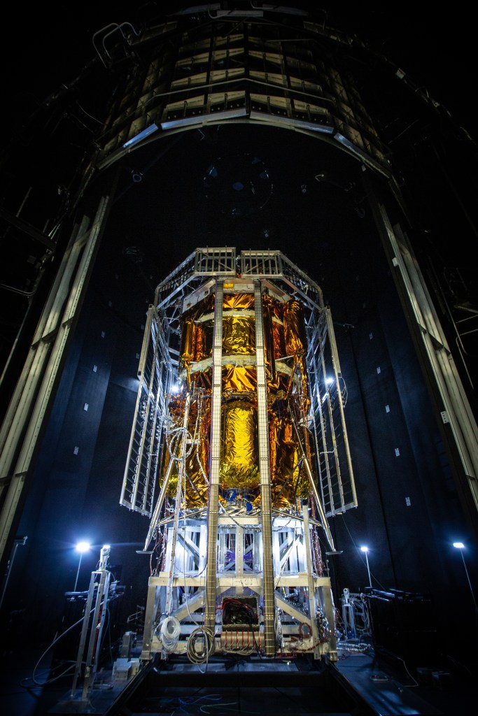 Blue Origin’s Blue Moon Mark 1 lunar lander, covered in gold thermal insulation, stands inside a dark testing chamber surrounded by support structures, cables, and lighting equipment.