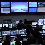 One large central screen, surrounded by four smaller screens on each side, spans the front wall of a control room displaying video and research flight data. Rows of engineers wearing headsets monitor real-time data on their computer screens.