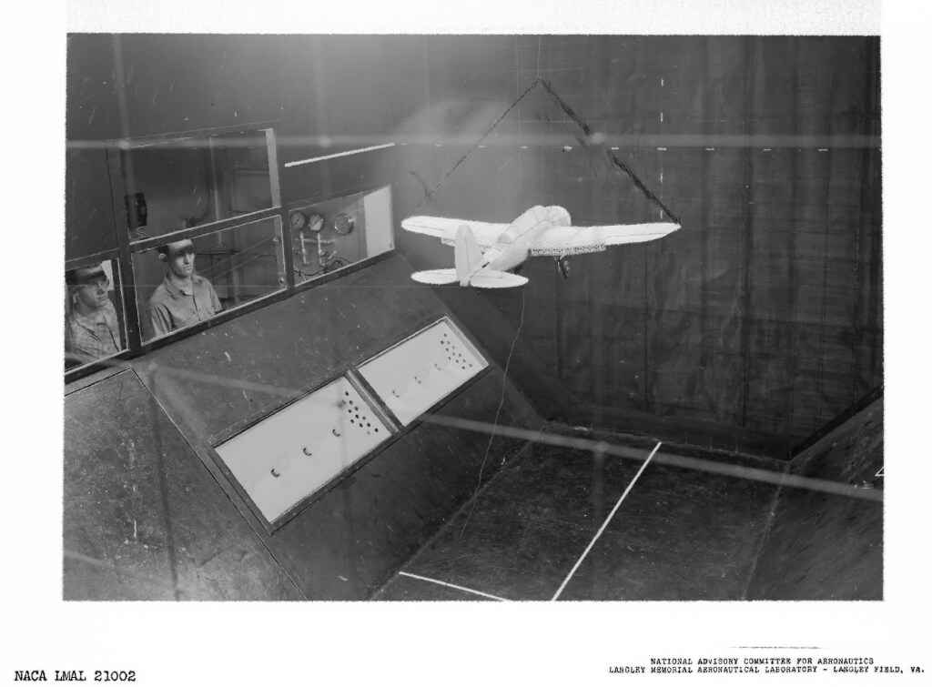 A light-colored aircraft model flies as two people watch from behind a window.