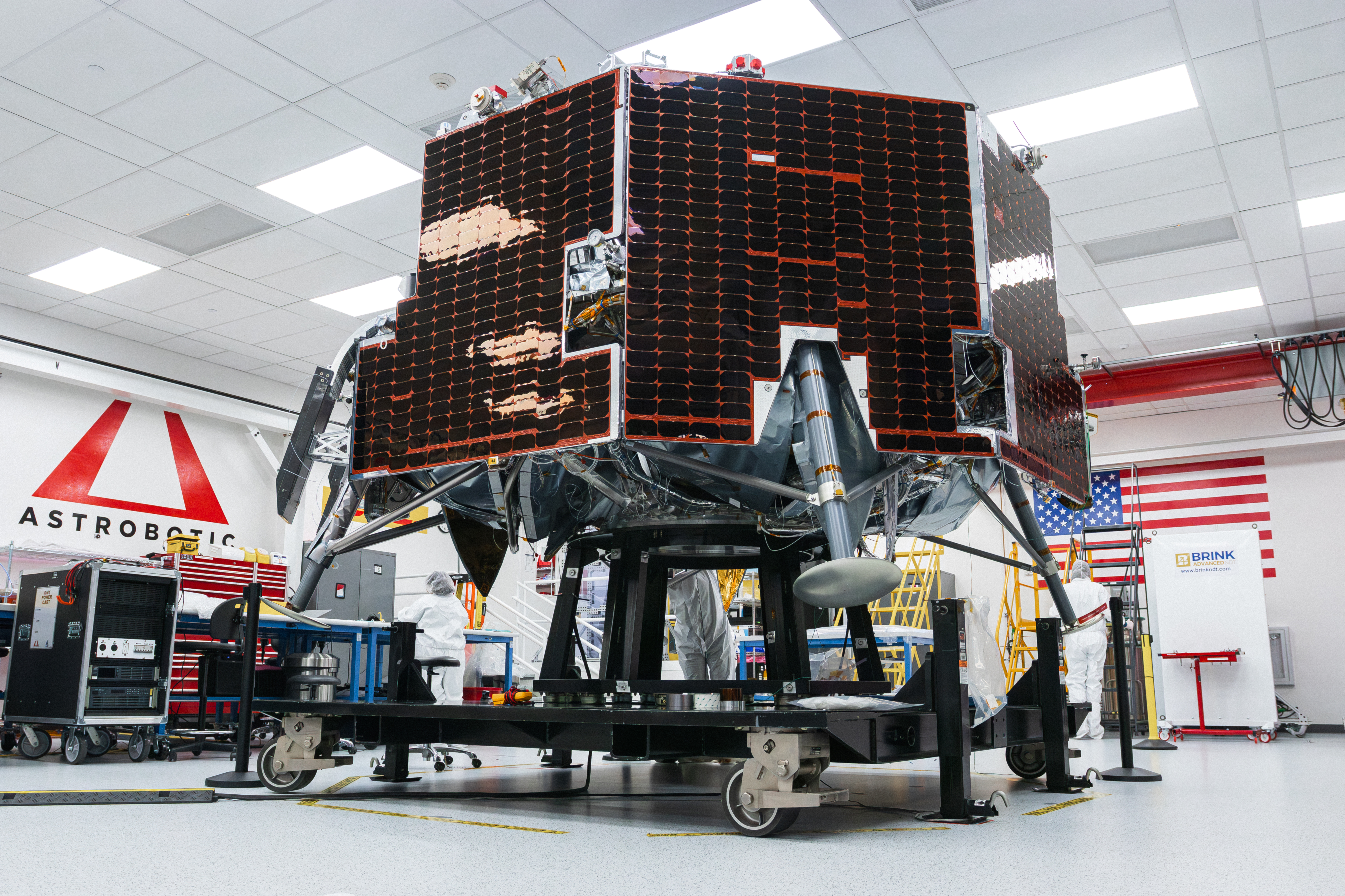 Astrobotic’s Griffin-1 lunar lander sits inside a clean room at the company’s facility. The lander is covered in dark solar panels and mounted on a wheeled test stand. Engineers in white clean-room attire work nearby, with the Astrobotic logo and a large American flag visible in the background.