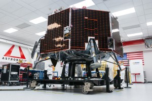 Astrobotic’s Griffin-1 lunar lander sits inside a clean room at the company’s facility. The lander is covered in dark solar panels and mounted on a wheeled test stand. Engineers in white clean-room attire work nearby, with the Astrobotic logo and a large American flag visible in the background.