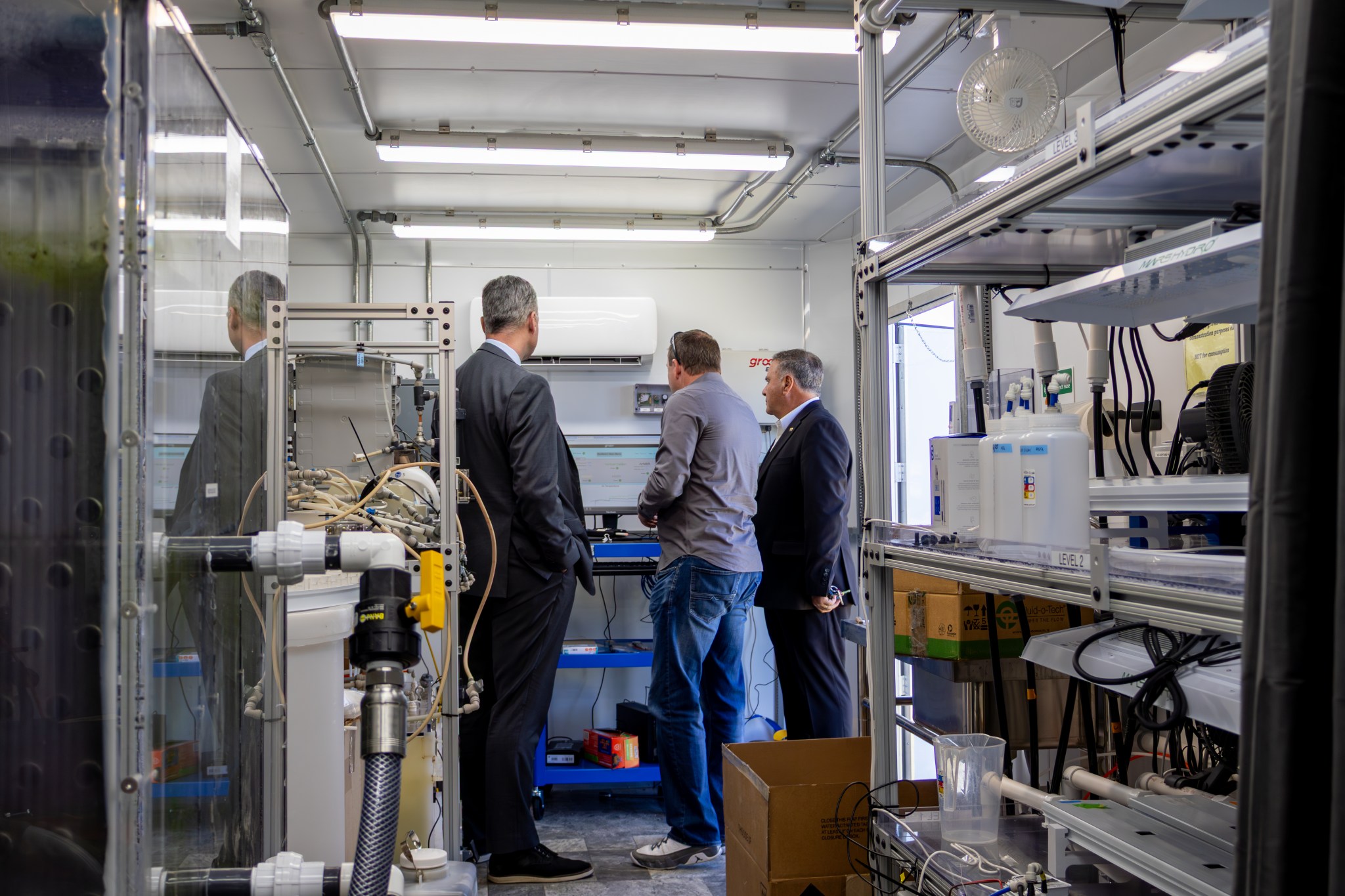 hree people stand inside a compact industrial workspace, examining equipment on a wall-mounted control panel. The room is filled with technical machinery, tubing, shelving, and containers used for laboratory or engineering operations. Bright overhead lights illuminate the space, and an open doorway is visible in the background.