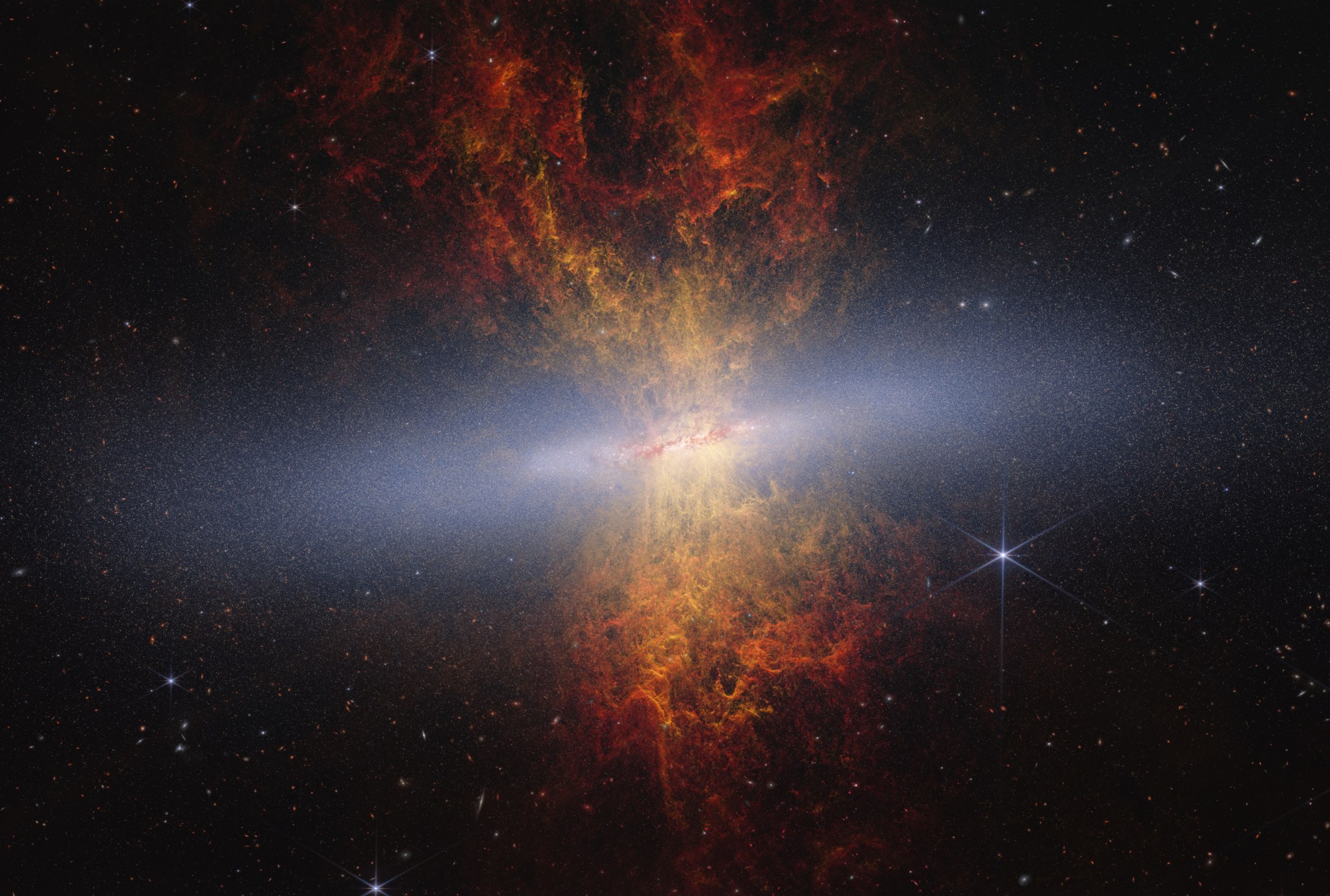 Hourglass-shaped plumes of gas shoot outward from above and below a bright disk-shaped center. The plumes are yellow near the galaxy’s bright center and gradually become redder as you move farther away. M82 is set against the black background of space, which has many distant galaxies that appear as small white and orange spirals, ovals, and points of light. On the right of M82 is a blue-white star with diffraction spikes.
