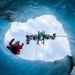 View from inside an ice cave looking up through an opening as a researcher in red gear and the EELS snake robot are lowered on cables against a cloudy sky, surrounded by blue glacial ice walls.