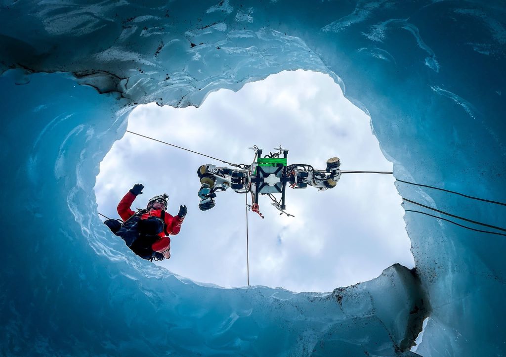 View from inside an ice cave looking up through an opening as a researcher in red gear and the EELS snake robot are lowered on cables against a cloudy sky, surrounded by blue glacial ice walls.