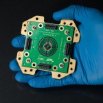 A gloved hand holds a green circuit board labeled NASA JPL Deep Space Optical Communications, featuring a central detector chip surrounded by radiating gold contacts and four edge connectors.