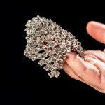 A hand holds a flexible sheet of 3D-printed metal fabric made of interlocking silver octahedral units, draped loosely across the palm against a black background.