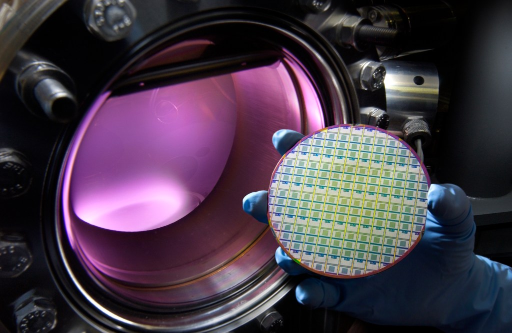 A gloved hand holds a circular semiconductor wafer printed with rows of multicolored microchip dies in front of an open vacuum chamber glowing purple.
