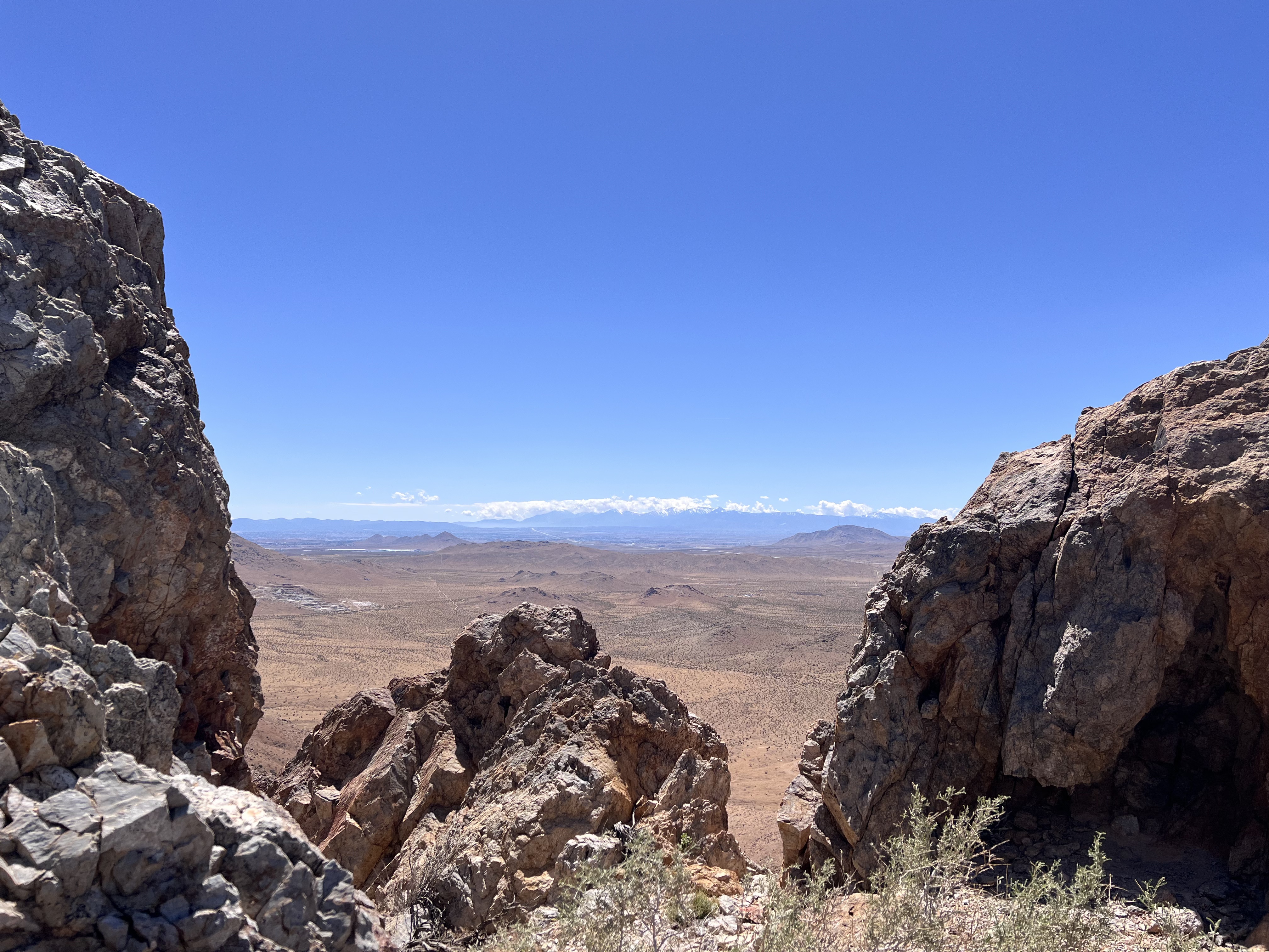 A wide-angle shot framing a vast desert plain viewed through a natural gap between jagged rock formations in the foreground. Distant snowcapped hills stretch across the far horizon beneath a thin line of cloud.