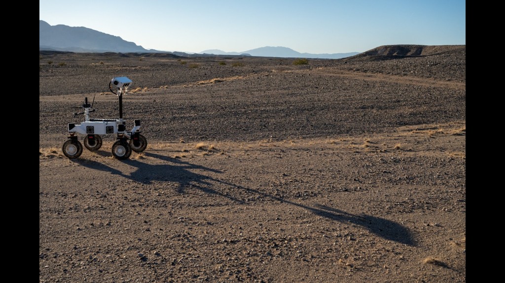 A white four-wheeled rover on the left side of the frame casts a long shadow across barren, brown ground toward the lower right. A few low, scrubby plants dot the desert landscape and mountains rise in the far distance under a pale blue sky.