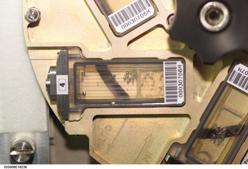 A close-up view of a mechanical fluid-handling component with a transparent chamber partially filled with liquid and small air bubbles. The chamber is mounted in a metallic frame with engraved flow-direction arrows and several barcode labels attached to surrounding parts.