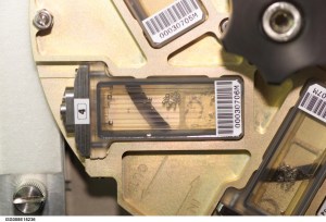 A close-up view of a mechanical fluid-handling component with a transparent chamber partially filled with liquid and small air bubbles. The chamber is mounted in a metallic frame with engraved flow-direction arrows and several barcode labels attached to surrounding parts.