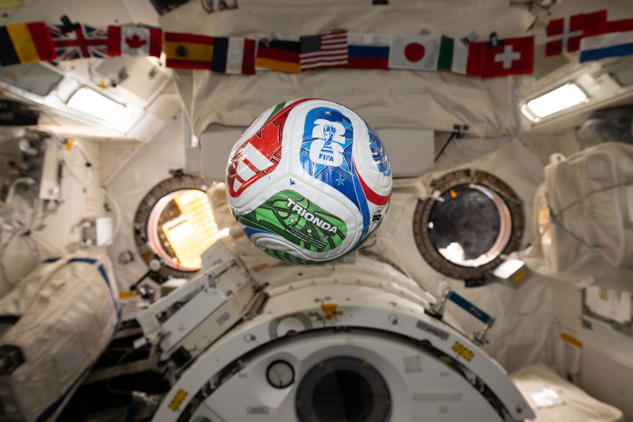 A white, red, blue, and green soccer ball floats inside the International Space Center. The FIFA logo is visible in the blue part of the ball facing the camera. The area in the background is mostly white, with a banner of country flags at the top of the photo.