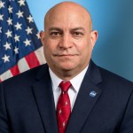 Jamie Dunn looks directly at the camera in his official portrait. He is wearing a dark suit with a red tie and a red and blue NASA meatball logo pin on his lapel. He has a slight smile. The portrait background is light blue. An American flag hangs over his right shoulder, on the left side of the image.