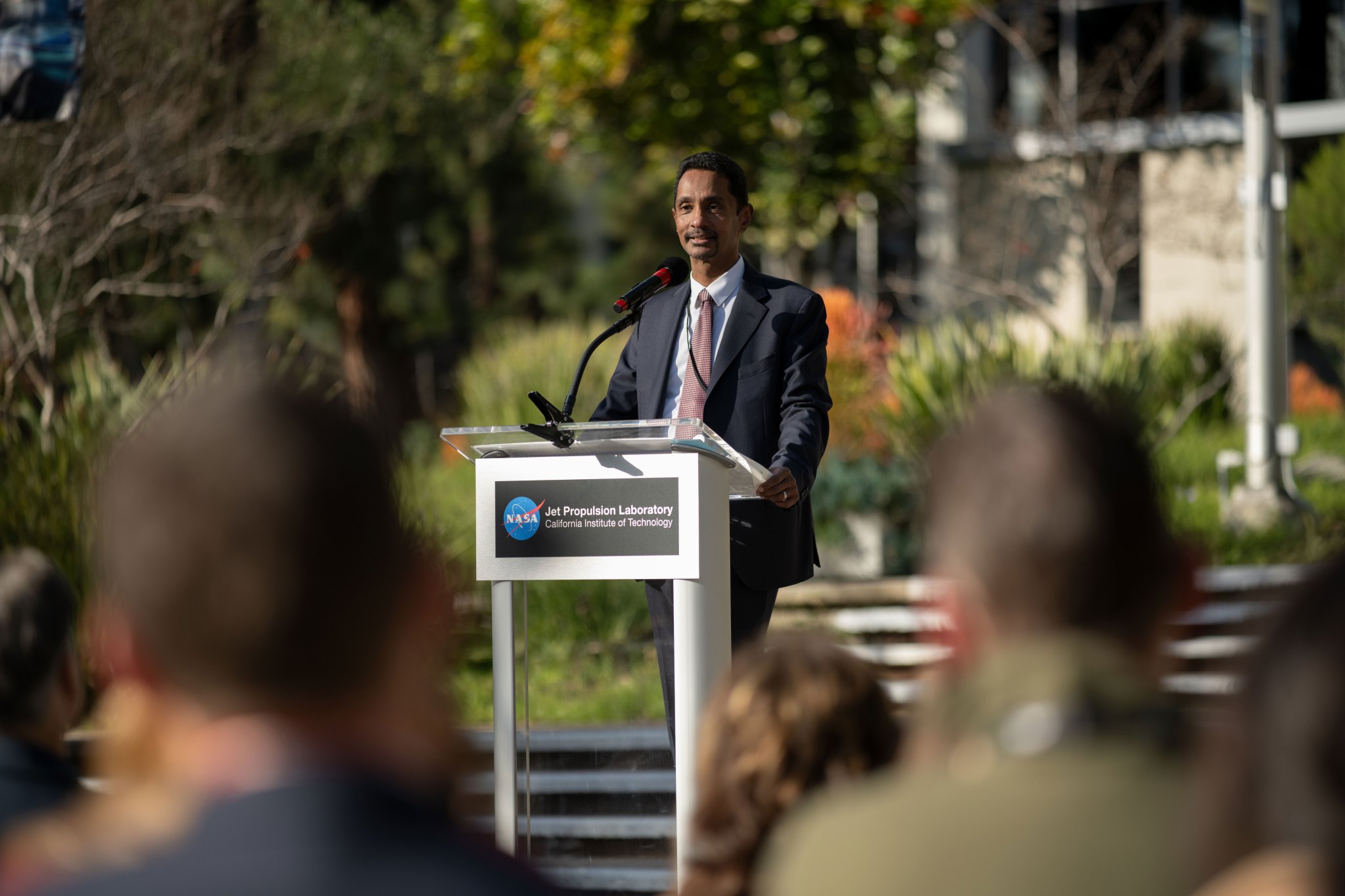 Wearing a dark suit, Caltech President Ray Jayawardhana speaks at a podium with a microphone outdoors, facing an audience. The podium displays the NASA Jet Propulsion Laboratory and California Institute of Technology logos.