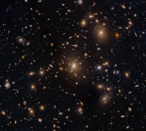 Numerous galaxies dot the scene and appear to cluster around the image center. The view includes large elliptical galaxies along with spiral and lenticular galaxies. Faint arcs of distant galaxies gravitationally lensed by the cluster appear in the upper-right quadrant of the image. A couple of foreground stars are also visible and easily distinguished by their diffraction spikes.