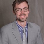 Headshot of a man with brown hair and scruff, wearing glasses, a gray suit and tie and blue striped shirt.