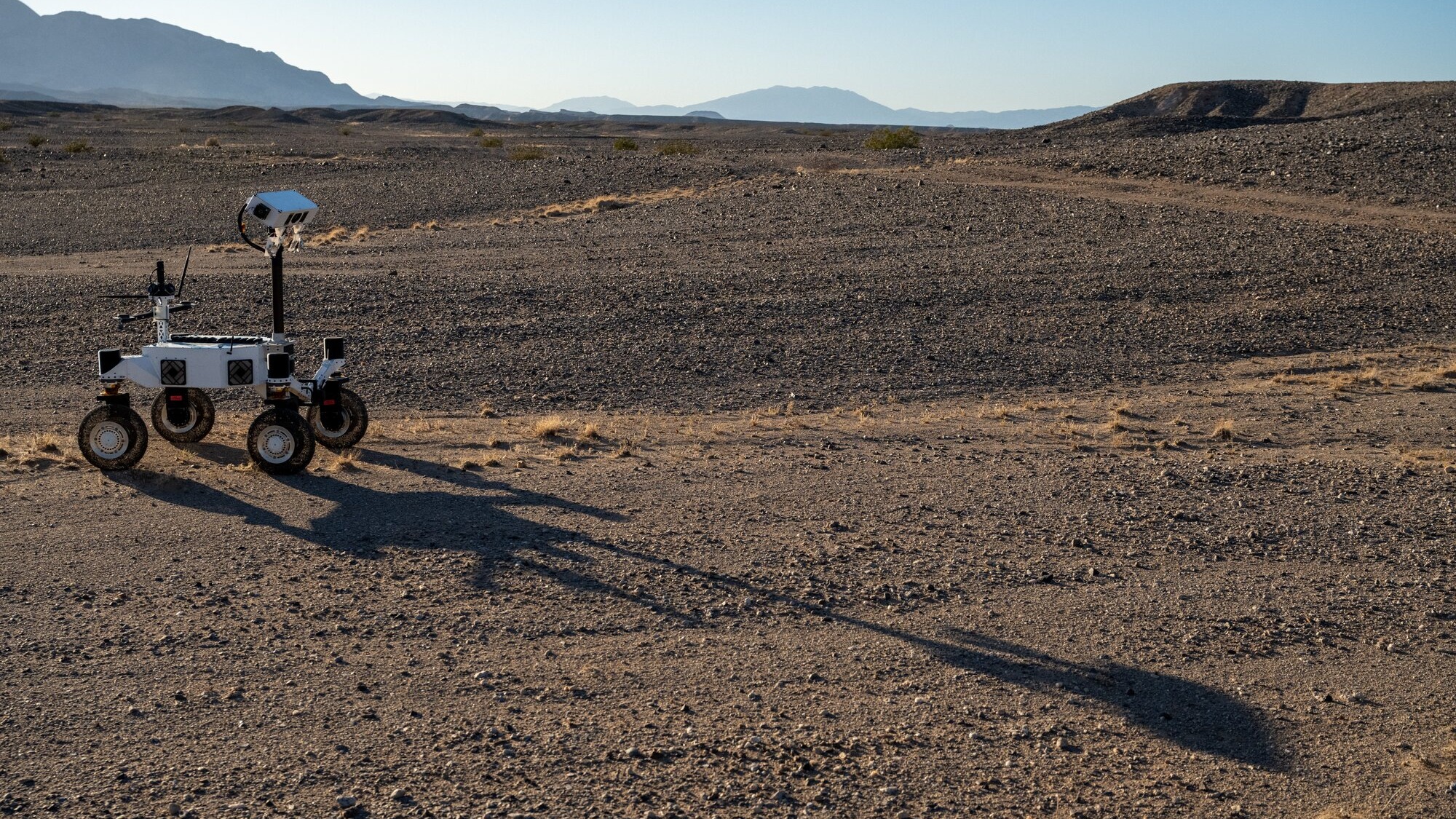 A white four-wheeled rover on the left side of the frame casts a long shadow across barren, brown ground toward the lower right. A few low, scrubby plants dot the desert landscape and mountains rise in the far distance under a pale blue sky.