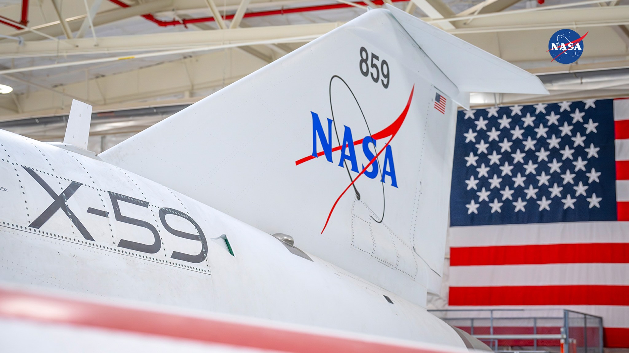 A closeup of NASA’s X59 tail section inside a hangar with the U.S. flag in the background.