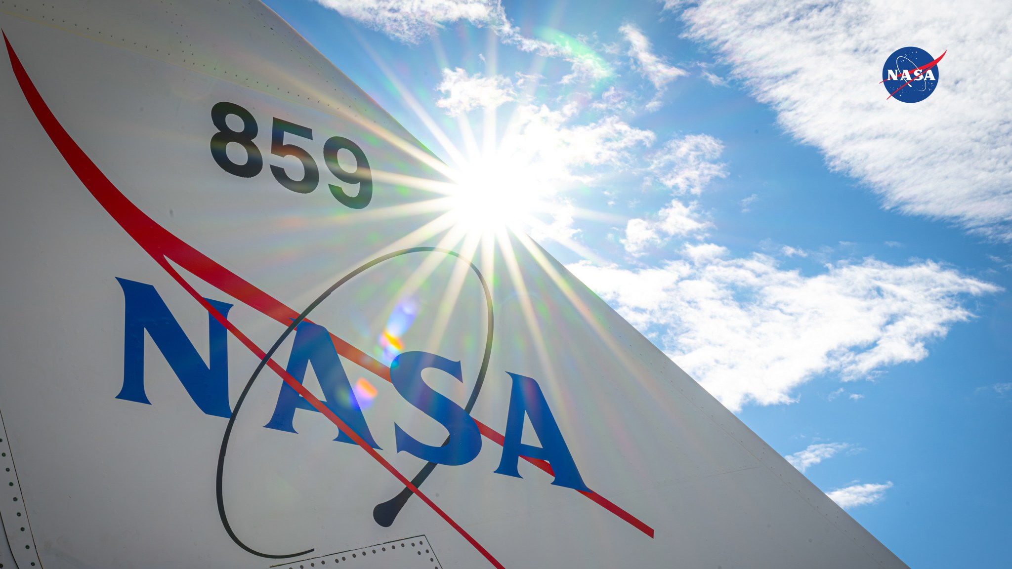 Sunlight shines across NASA’s X-59 tail section, showing the aircraft number and NASA logo against a partly cloudy sky.