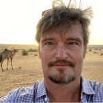 Headshot of a man with light brown hair and goatee, taken selfie style with a camel visible over his right shoulder.